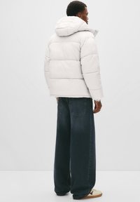 Man in een witte gewatteerde jas met capuchon, met een brede pijpen donkere spijkerbroek en witte sneakers met rode accenten, staande tegen een eenvoudige achtergrond.