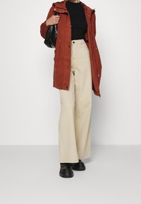 Chaqueta exterior roja con capucha, pantalones beige de pierna ancha y botas negras gruesas. La chaqueta cuenta con cremalleras y botones de presión, con un acabado texturizado.