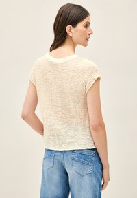 Licht beige T-shirt met korte mouwen, met een gekreukt textuur, ronde halslijn en een relaxte pasvorm. G gedragen met blauwe high-waisted denim jeans.
