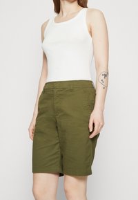 Shorts vert olive à coupe droite avec poches latérales, associés à un débardeur blanc ajusté. Tissu en coton avec une texture lisse.