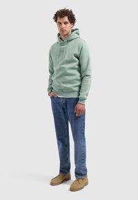 Jonge man die staat, gekleed in een lichtgroene hoodie, blauwe spijkerbroek en tan schoenen tegen een effen lichtgrijze achtergrond.