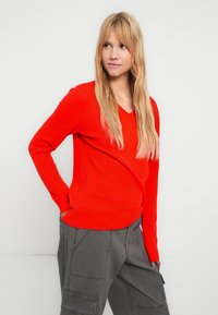 Κόκκινο πουλόβερ με λαιμόκοψη σε σχήμα V, με ριμπ μανσέτες και τελείωμα, φτιαγμένο από μαλακό υλικό. Φοράει πάνω από γκρι παντελόνι cargo, παρουσιάζεται σε ουδέτερο φόντο.