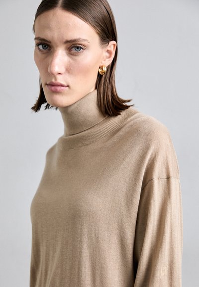 Beige turtleneck sweater with a ribbed collar and long sleeves; features a subtle texture and a small embroidered logo on the sleeve.