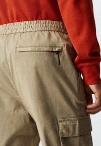 Beige cargobroek met een elastische tailleband, voorzien van een ritsafsluitbare achterzak en een zijvak. De stof ziet er soepel en lichtgewicht uit.