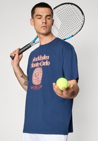 Marineblå t-shirt med rød grafik, der har teksten "Stockholm Monte Carlo Tennis Club." Modellen holder et tennisbat og en bold.