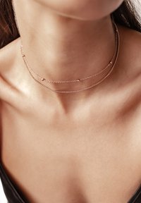 Trzy delikatne łańcuszki w kolorze różowego złota z małymi koralikami, ułożone na szyi, na tle gładkiej karnacji i ciemnego materiału.