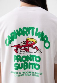 T-shirt bianca in cotone con una stampa grafica colorata sul retro. Il design include testo verde e un personaggio dei cartoni animati rosso che corre.