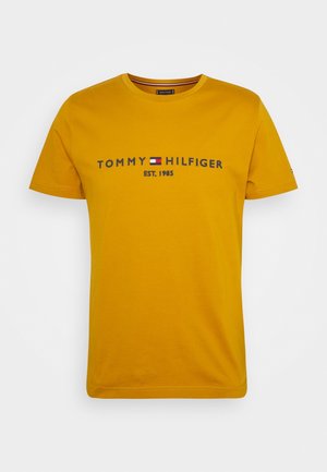 Senfgelbes T-Shirt mit kurzen Ärmeln mit dem Schriftzug „Tommy Hilfiger EST. 1985“ und einem kleinen rot-weiß-blauen Flaggenlogo in der Mitte der Brust.