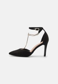 Scarpa nera in suede con tacco alto e punta affilata, cinturino alla caviglia e dettagli in catena argentata lungo la parte superiore. Fodera interna morbida e tacco sottile.
