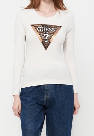 Mujer con camiseta blanca de manga larga con gran logotipo metálico en oro rosa y estampado de leopardo de Guess y vaqueros azules.