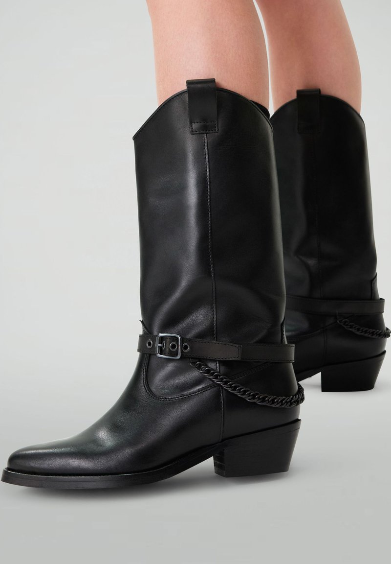 Bottes de cowboy en cuir noir avec un bout pointu, hauteur mi-mollet, détail de sangle à la cheville et talon épais bas. Texture lisse avec une accent enchaîné.