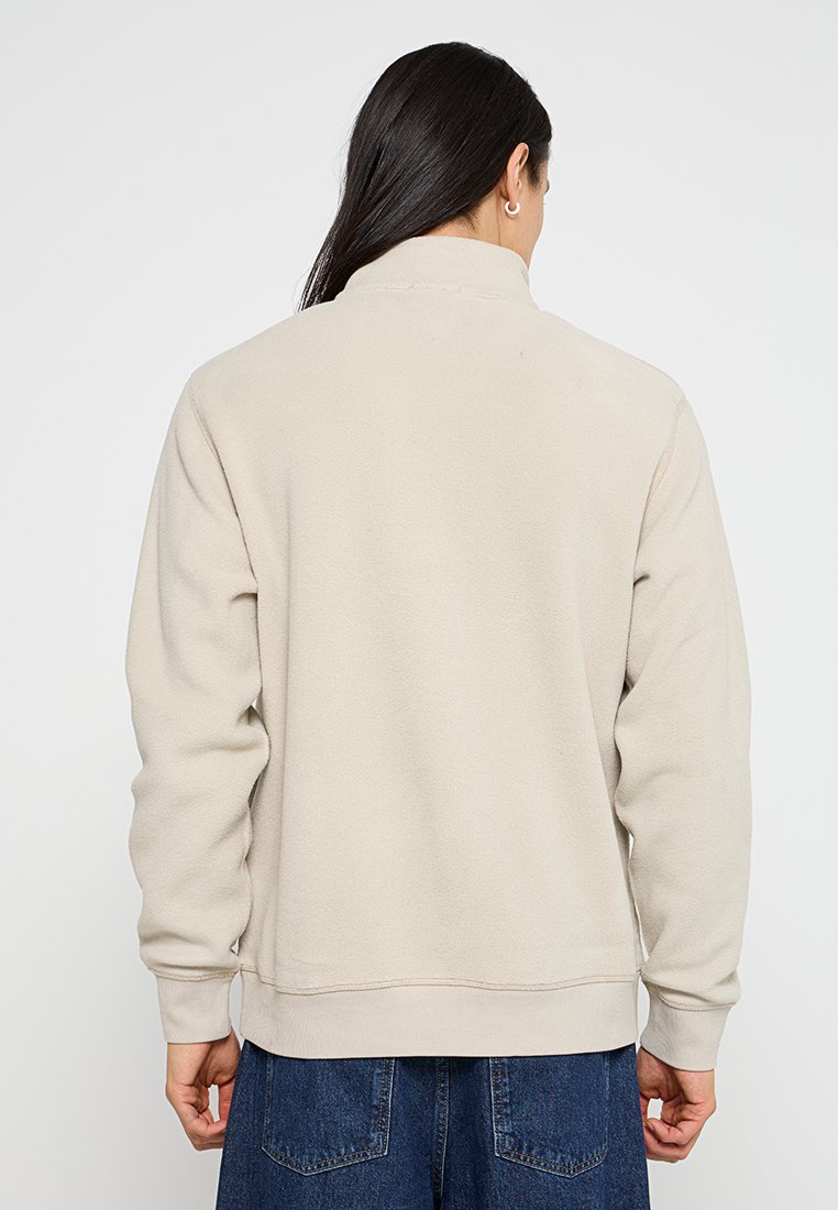 Felpa in fleece beige con collo alto, polsini e orlo a coste. Texture liscia, vestibilità casual e senza loghi o motivi visibili.