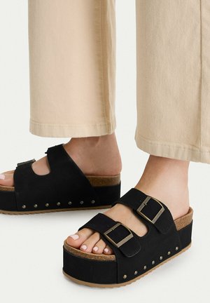 Feet in beige pants wearing black platform sandals with two buckle straps and stud details along the sole edge on a white background.