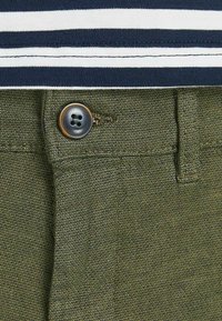 Pantaloni verde oliva con tessuto testurizzato, chiusura con bottone e passanti per cintura. Abbinati a una maglietta a righe blu e bianca.