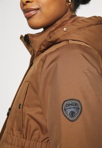 Chaqueta de plumas marrón con capucha, botones de presión y un parche con el logo "ONLY" en la manga. Tela suave y diseño estructurado.