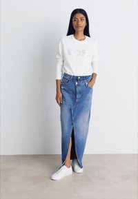 Denim maksi krilo s sprednjim razporkom, svetlo modre barve, s strapcem na robu, v kombinaciji z belim puloverjem z zlatim logotipom in belimi supergami.