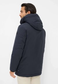 Uomo che indossa un giubbotto invernale con cappuccio di un blu scuro, rivolto a sinistra con schiena e lato visibili, su uno sfondo bianco uniforme.