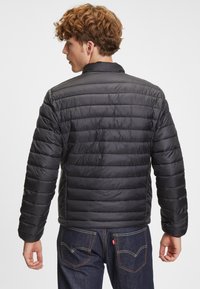 Černá péřová bunda s horizontálním prošívaným vzorem, stojáčkovým límcem a hladkým povrchem. Nosená přes tmavé džíny, zobrazuje zadní pohled.