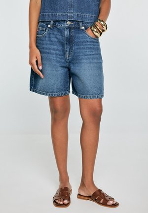 Niebieskie, średniej długości szorty dżinsowe noszone z brązowymi, płaskimi sandałami i złotymi bransoletkami na jednym nadgarstku, modelka stoi z rękami swobodnie ułożonymi, jedną w kieszeni.