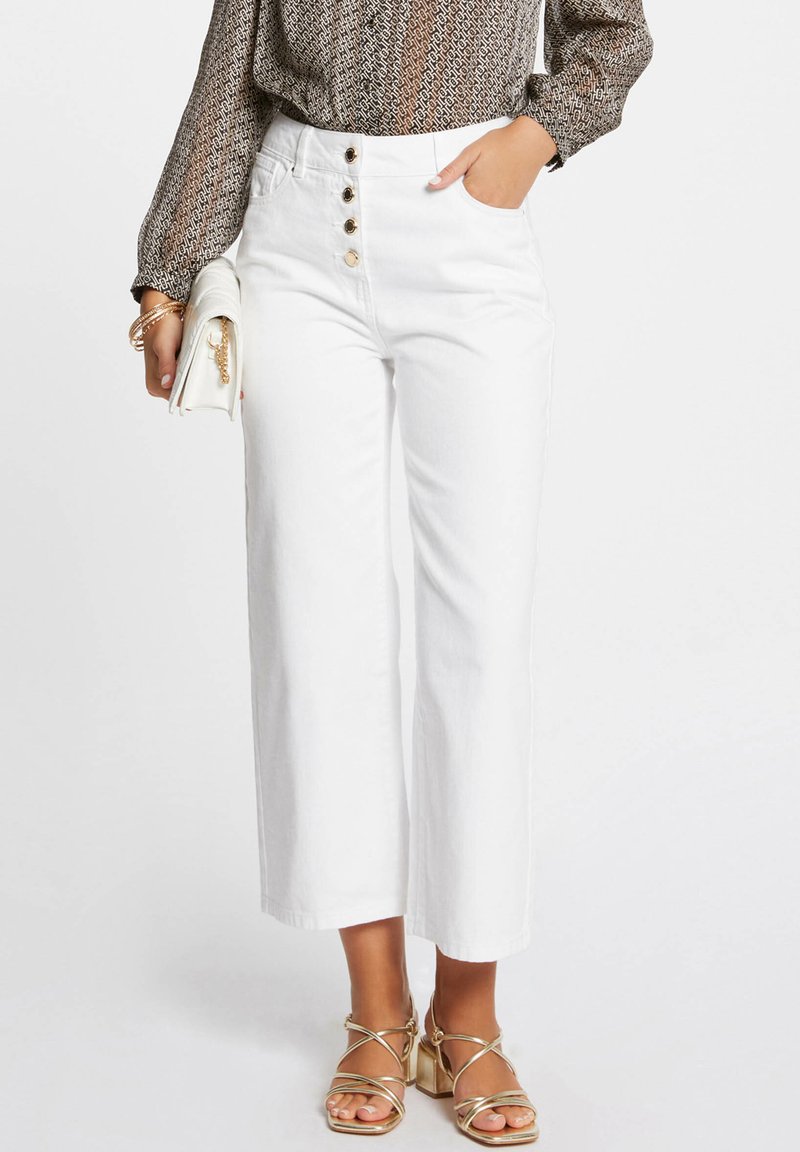 Pantalones blancos de cintura alta con botones frontales, pernera ancha y bolsillos laterales. Combinados con tacones dorados con tiras y un bolso de mano blanco. Blusa texturada en la parte superior.