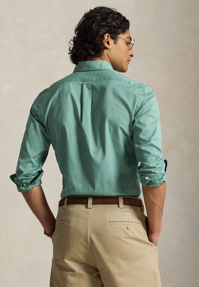 Homme aux cheveux bouclés foncés portant des lunettes, une chemise verte retroussée, un pantalon beige et une ceinture marron, debout les mains dans les poches.