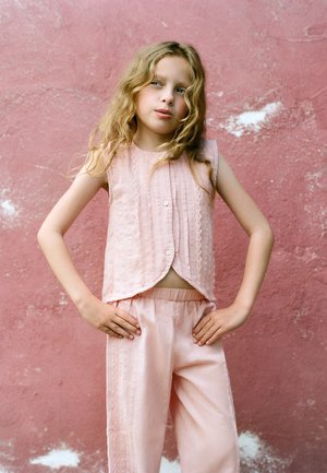 Jong meisje met lang golvend blond haar staat met handen in de zij, draagt een roze mouwloos topje en bijpassende broek voor een gestructureerde roze muur.