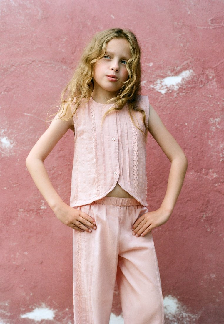 Jeune fille aux longs cheveux blonds ondulés, debout, mains sur les hanches, portant un haut rose sans manches et un pantalon assorti devant un mur rose texturé.