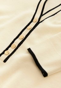 Gilet crème avec bord noir, comportant six boutons dorés brillants sur le devant et une texture douce sur tout le tissu.