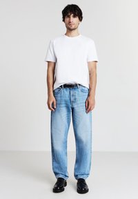 Uomo in piedi contro uno sfondo neutro, indossa una maglietta bianca, jeans azzurri chiari con cintura nera e scarpe nere, volto in avanti con le braccia rilassate.