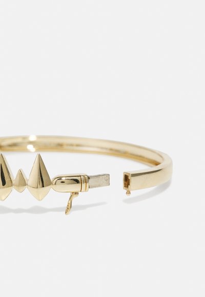 Kuldtoonist metallist bangle-käevõru, millel on viis teravaotstega niiti. Kindel klamber ja sile, ümar riba läikiva viimistlusega.