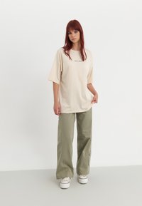 T-shirt beige oversize à manches courtes avec un logo sur la poitrine, associé à un pantalon large vert olive et des baskets blanches.