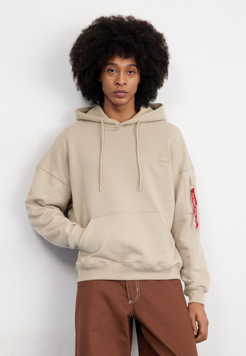 Felpa beige con cappuccio, tasca frontale, cordino e un accento rosso sulla manica sinistra che presenta una tasca con zip.
