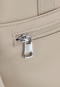 Borsa in tessuto beige con zip argentata e linguetta. Presenta dettagli cuciti e una superficie liscia. Il logo è impresso sulla linguetta della zip.