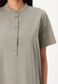 Svetlo siva majica s kratkimi rokavi in stoječim ovratnikom, z gumbasto zadrgo in subtilno teksturo.