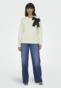 Felpa color avorio con maniche lunghe, caratterizzata da un grande fiocco nero accentato sulla spalla sinistra, abbinata a jeans a gamba larga blu.