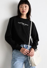 Sudadera negra con el logotipo de "Calvin Klein Jeans", combinada con jeans de mezclilla azul claro. Se sostiene un bolso blanco con correa ajustable en una mano.