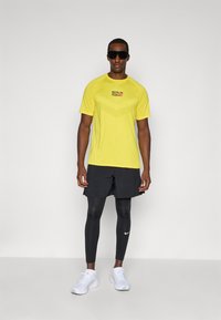 Menns gule treningsskjorte med teksturert design, "BERLIN RUN" logo. Parret med svarte shortser og leggings, hvite joggesko og solbriller.