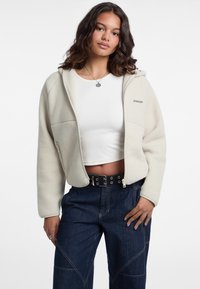 Giacca in fleece color crema con cappuccio, dotata di una cerniera frontale completa e due tasche laterali. Indossata sopra un top corto bianco e jeans blu.