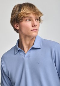 Ljusblå polo skjorta gjord av texturerad tyg, med v-ringad hals och två knappar i placket, samt en liten broderad logotyp på bröstet.