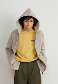Beige zip-up hoodie med huva över en gul tröja. Modellen har på sig olivgröna byxor. Tyget verkar mjukt med en avslappnad passform.