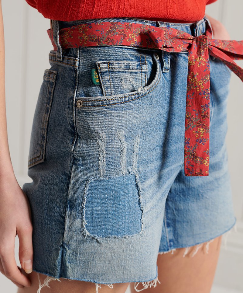 Jeansshorts mit fransigem Saum, aus hellem blauen Stoff, mit abgenutzten Stellen und einem roten Blumengürtel, der in der Taille gebunden ist.