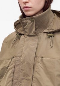 Femme portant une veste beige à col montant avec poches zippées et cordons réglables à la capuche, sur fond blanc uni.