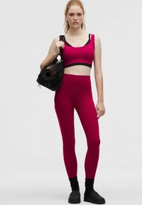 Magenta sportmelltartó fekete pántokkal, ami egy összeillő magas derekú leggingsszel van párosítva. Fekete táska, amiben egy feltekert jógaszőnyeg van a vállon.