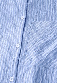 Light blue striped shirt featuring a pocket on the left side, made of textured fabric with white buttons and a wrinkled appearance.