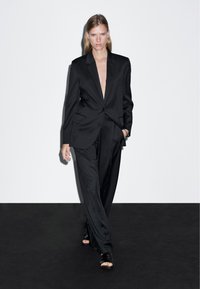 Czarny garnitur klasyczny z głębokim dekoltem w kształcie litery V i pojedynczym zapięciem na guzik. Gładki materiał, proste spodnie, oraz sandały na cienkich paskach w kolorze czarnym.