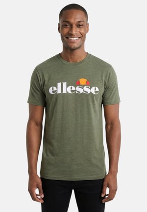 Homme souriant portant un t-shirt ellesse vert olive et un pantalon noir, debout devant un fond blanc uni.