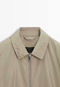 Béžová lehká bunda s špičatým límcem a stříbrným předním zipem, zobrazená od hrudi nahoru na bílém pozadí.