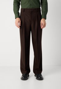 Pantalons habillés marron avec une coupe ample, des plis marqués, un devant plat et des poches latérales, associés à des chaussures noires.