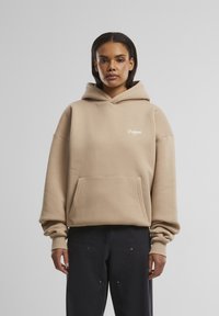 Beige hettegenser med kengurulomme og ribbestrikkede mansjetter. Har hvit logo tekst på brystet. Myk fabric og avslappet passform.