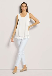 Weißes ärmelloses Oberteil mit einem runden Ausschnitt und Spitzenbesatz am Saum. Kombiniert mit hellblauen Jeans und beigen Ballerinas. Glatte Stofftextur.